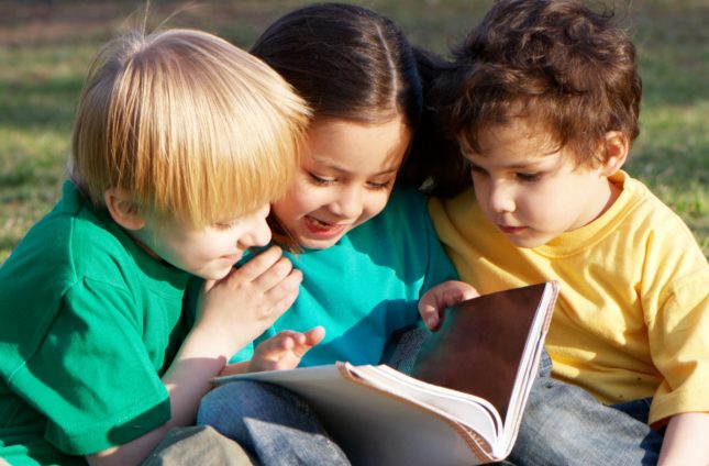 A girl and two boys sitting on the grass, reading a book